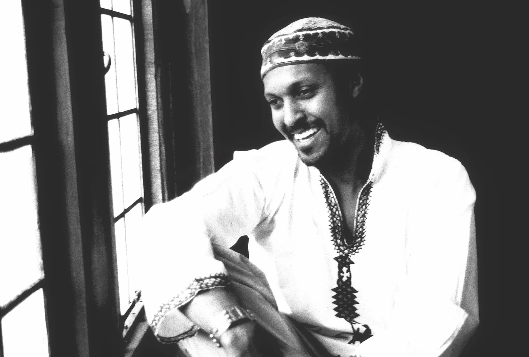 Person wearing embroidered traditional clothing and a patterned cap sits beside a window in a black-and-white portrait.