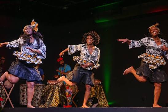 Three barefoot dancers in coordinated patterned costumes perform on stage, balancing on one leg with outstretched arms, while musicians play in the background.